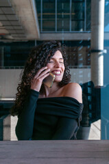 Joyful woman with curly hair laughing while talking on smartphone outdoors