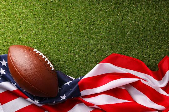 Football rests on green grass beside the American flag - Powered by Adobe