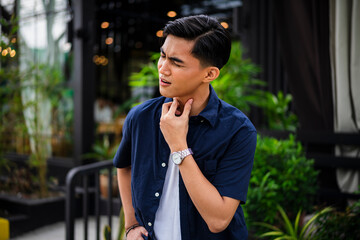 The young Asian man tilts his head and touches his neck, showing discomfort or pain while standing in the vibrant outdoor café setting.