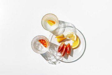 Summer still life scene with clear glass cups and plate. Glittering glasses of water with slices of fruit on white table at sunlight. Long harsh shadows and caustics. Summer vacation concept, above