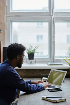 African office worker in eyeglasses sitting at the table and typing on laptop in front of the window at office