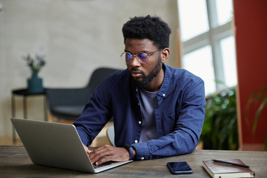 Horizontal image of African young businessman siting at his workplace and using laptop, he concentrating on his online work