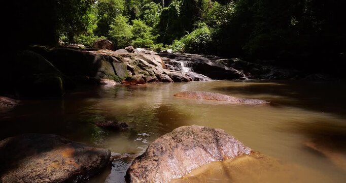 Sunlight filters through lush canopy illuminating cascading water flowing over rocks in brown pond, tilt down shot, large stone on foreground. Lubuk Teja waterfall hidden in tropical rainforest