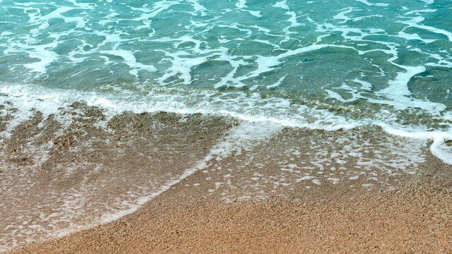 the clear, turquoise water of the ocean gently meets a light golden sandy beach, with soft waves creating white foam at the shoreline. - Powered by Adobe