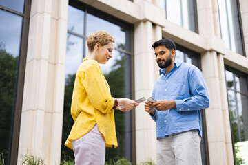 Obraz premium A woman in a yellow shirt hands money to a man in a blue shirt, both outdoors in front of a building.