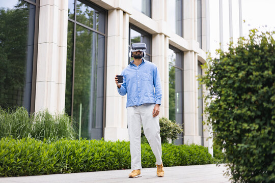 A man wearing a VR headset holds a coffee cup while walking outside a modern building with green hedges.