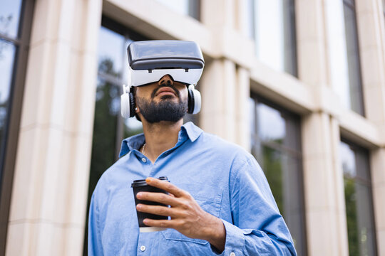 A man wearing a VR headset drinks coffee outdoors, experiencing virtual reality in an urban setting. - Powered by Adobe
