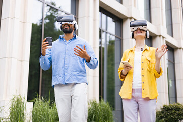 A diverse couple experiences virtual reality outdoors. They wear VR headsets and interact with their surroundings. The man holds a coffee cup.