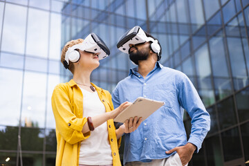 A diverse couple explores virtual reality using VR headsets and a tablet in front of a modern office building.