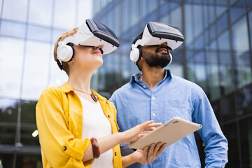 A diverse couple wearing VR headsets explore augmented reality with a tablet outdoors near a modern building.
