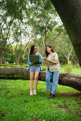 Fototapeta premium Two Young Women Collaborating Outdoors While Using Laptop and Reading Notes in a Lush Green Park Surrounded by Trees and Nature