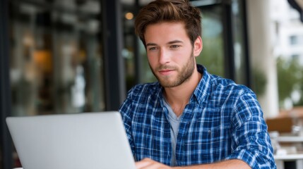Fototapeta premium Young man in blue checkered shirt working on laptop in coffee shop, focused facial expression, natural daylight, modern urban style 