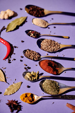 Vertical image of large number of spoons lying in a row with different spice with seeds around them isolated on purple background