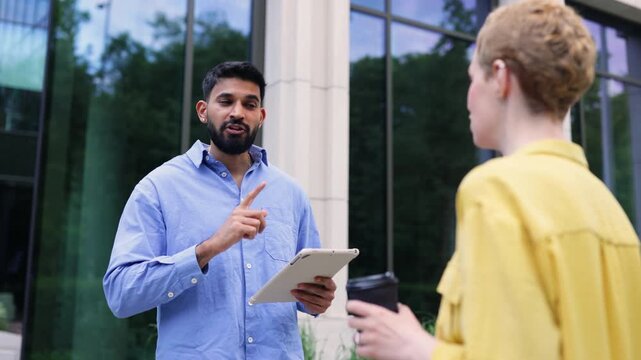 A man in a blue shirt converses with a person in a yellow shirt outdoors, holding a tablet and coffee, against a backdrop of a building.