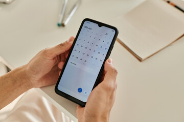 Horizontal close-up image of man sitting at the table and holding mobile phone, he opening calendar and looking at dates