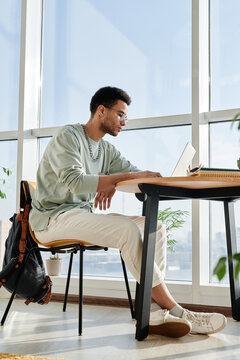 Vertical image of young man sitting at desk and studying online using his laptop in the room with big windows