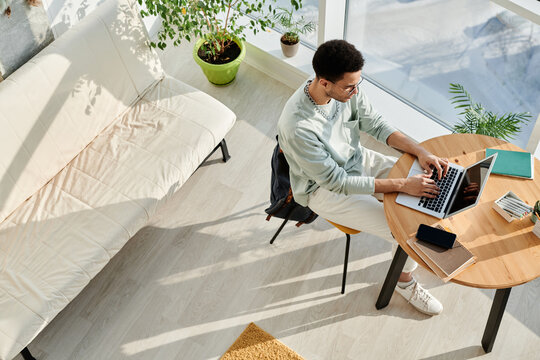 High angle view of young businessman sitting at the table typing on laptop and working online at modern office with sofa and big windows - Powered by Adobe