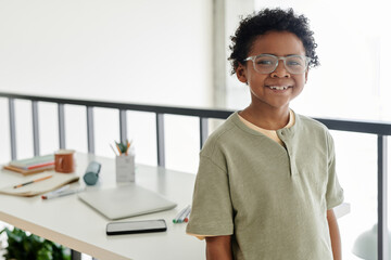 Horizontal portrait of African schoolboy smiling at camera while standing in the room with desk with books in the background