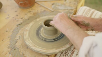 A close-up shot captures the skilled hands of a potter shaping wet clay into a pot on a spinning throwing wheel. The artisan creates handmade ceramic ware in a rustic workshop.