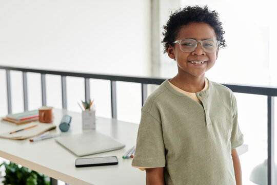 Horizontal portrait of African schoolboy smiling at camera while standing in the room with desk with books in the background - Powered by Adobe