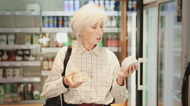 Mature woman chooses natural cheese in a store with organic products. Pensioner buys farm cheeses in supermarket for healthy eating. High quality 4k footage