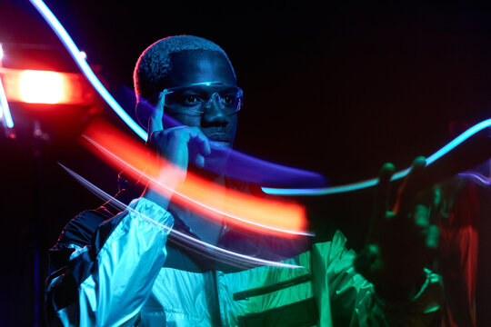 Horizontal medium portrait of African American guy with blond hair wearing stylish outfit and transparent eyeglasses standing in dark studio room in neon lights using AR technology