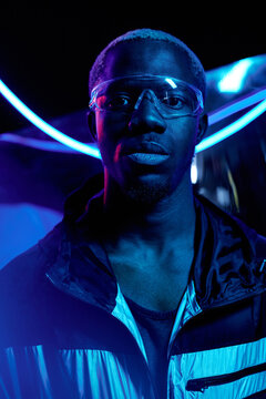 Vertical studio portrait of African American guy with blond hair wearing stylish outfit and futuristic transparent eyewear standing in dark room in neon lights looking at camera