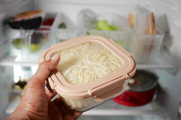 Hand holding container of noodles inside a refrigerator
