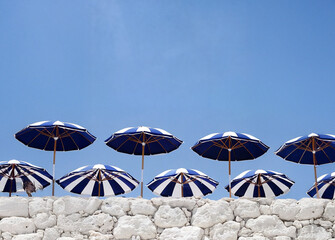 A series of blue and white striped umbrellas arranged on a white stone wall against a clear blue sky.