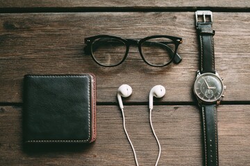 Flatlay of black wallet, glasses, watch, and headphones on a dark wooden surface