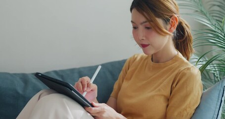 Positive young Asia female freelance casual wear use tablet remote working sit on sofa in living room at home. Work from house, financial plan, digital nomad job and Gen Z talent concept. - Powered by Adobe