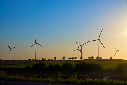 Wind turbines in open farmland with green fields and trees under evening sky. Windmills for production clean energy at sunset. Concept of renewable energy, sustainable resources and green technology