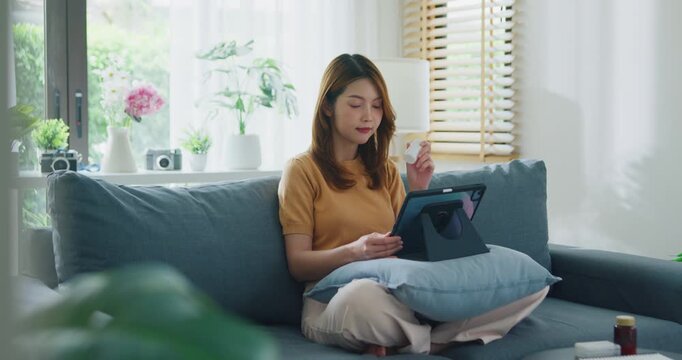 Sick young Asian woman hold medicine sit on couch video call with digital tablet consult with doctor in living room at home. Girl take medicine after doctor order, telemedicine, mental health concept.