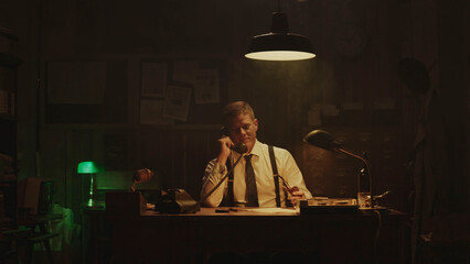 Focused detective working late at night in his office, taking notes while on the phone