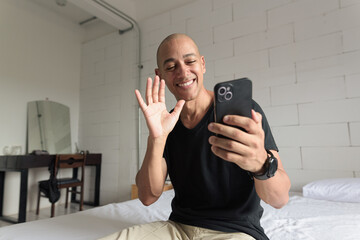 Bald Hispanic man sitting on bed at home using phone having video call waving hand