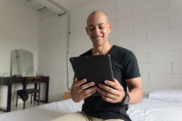 Bald Hispanic man sitting on bed at home using digital tablet computer