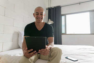 Hispanic bald happy man sitting on bed while using digital tablet computer © Ranta Images
