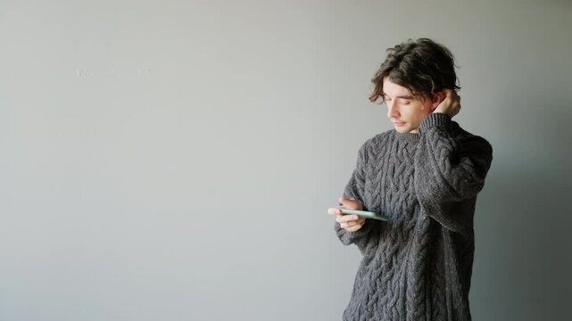 Medium slowmo portrait of handsome young Caucasian man with curly brown hair in graphite sweater scrolling on smartphone standing against clear grey background