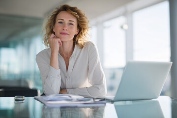 Confident businesswoman sitting at office desk with laptop and documents, smiling and looking at camera