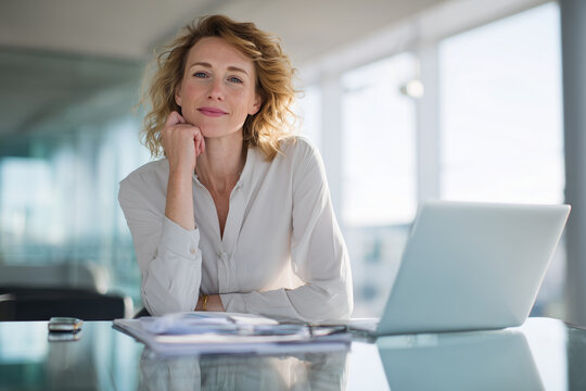 Confident businesswoman sitting at office desk with laptop and documents, smiling and looking at camera
