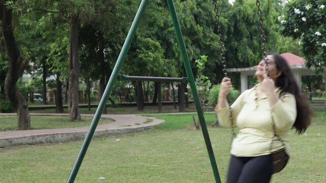 Mature Indian woman riding swing outdoor in summer park. Brunette Female swinging and enjoying carefree. 4k