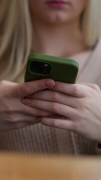 Close-up of young lady operating iPhone with green case while seated at wooden table, her hands are focused on screen, showing attention to detail in a relaxed environment
