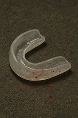 Clear mouthguard with blood stains lying on a dark floor, suggesting intense physical impact or a violent moment in a combat sport.