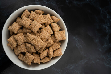 A white bowl filled with crunchy, whole grain cereal squares sits on a dark countertop, highlighted by natural morning light. Perfect for starting the day right.