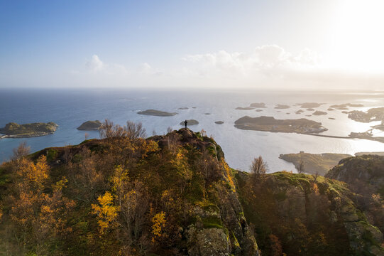 Aerial view of a lone figure atop a rugged, golden-hued peak overlooking the serene, island-dotted waters of Henningsv&Atilde;&brvbar;r, Nordland, Norway.