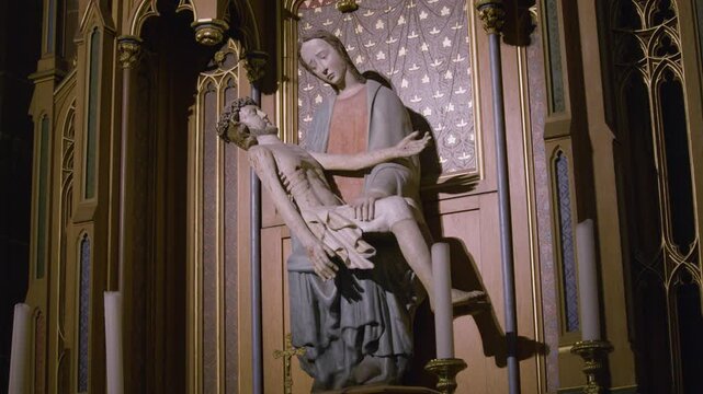 Stone religious statue of Mary and Christ inside a gothic church with wooden altar and candles. Tilt shot of sculpture composition:  the Virgin Mary holding the body of Christ