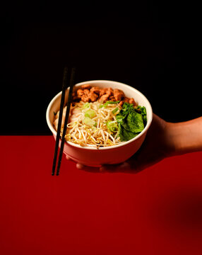 Hand holding a bowl of Indonesian chicken noodles ( mie ayam )  with minced chicken, bean sprouts, greens, and chopsticks on a bold red-black background. Great for menu, delivery, and branding.