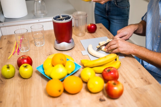 Close up of adult couple preparing smoothie in their kitchen. They are mixing various fruits in electric juicer. Man is slicing banana.