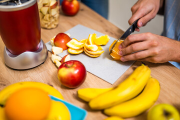 Close up of adult couple preparing smoothie in their kitchen. They are mixing various fruits in electric juicer. Man is slicing orange.