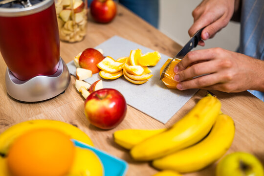 Close up of adult couple preparing smoothie in their kitchen. They are mixing various fruits in electric juicer. Man is slicing orange. - Powered by Adobe